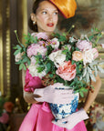 Woman looking away, wearing light pink day gloves holding flower vase.