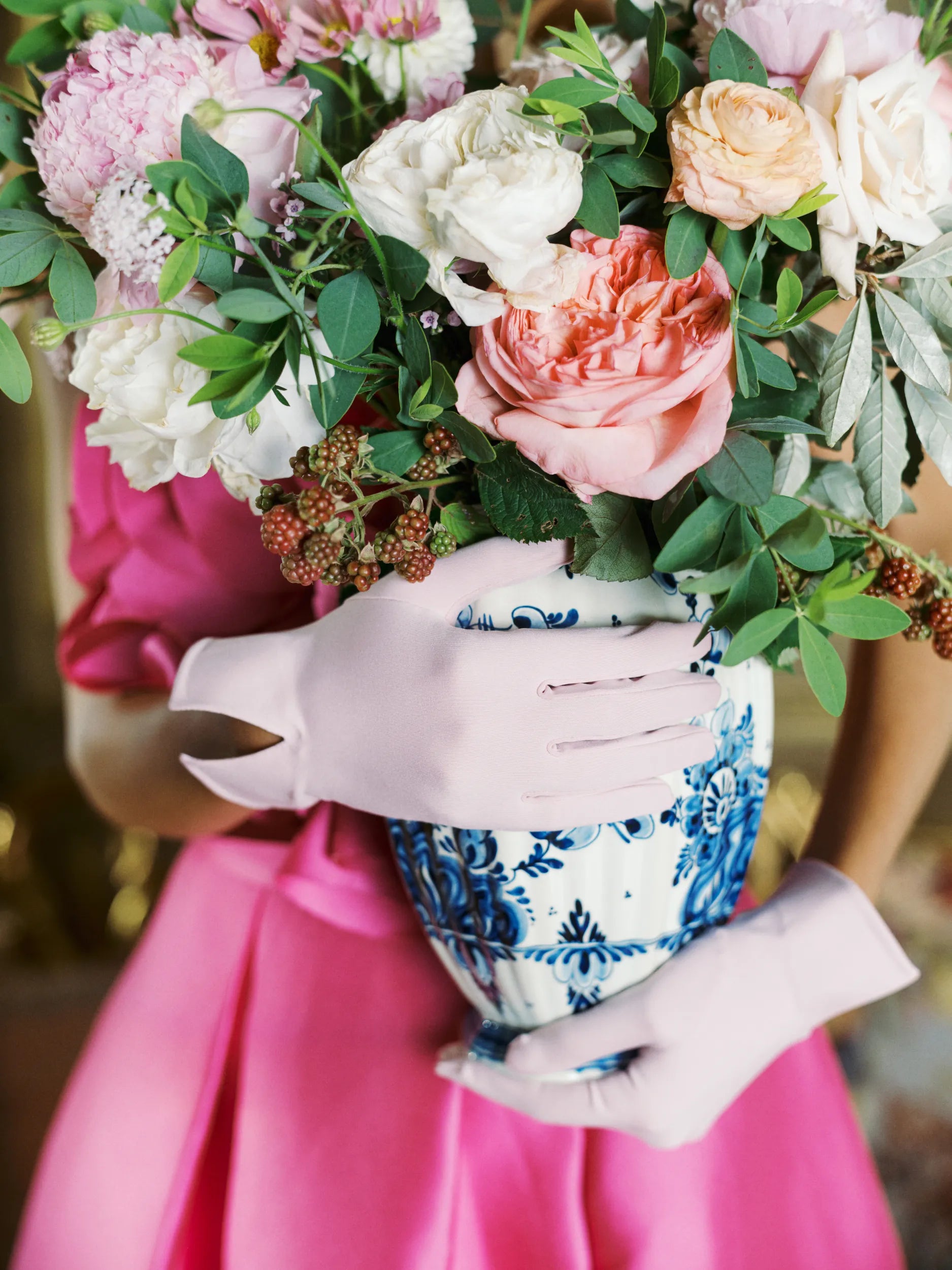 Woman wearing light pink wrist gloves holding large vase of flowers.