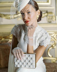 Woman in wedding dress & wedding hat wearing tulle white gloves holding white satin clutch.