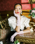 Woman sits, poised, wearing pink, elbow length gloves.