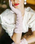 A glamorous woman rests her head in her hand, wearing pink elbow, long gloves.