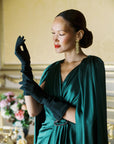 A woman fixing her black mid length Jill gloves in a formal green dress.