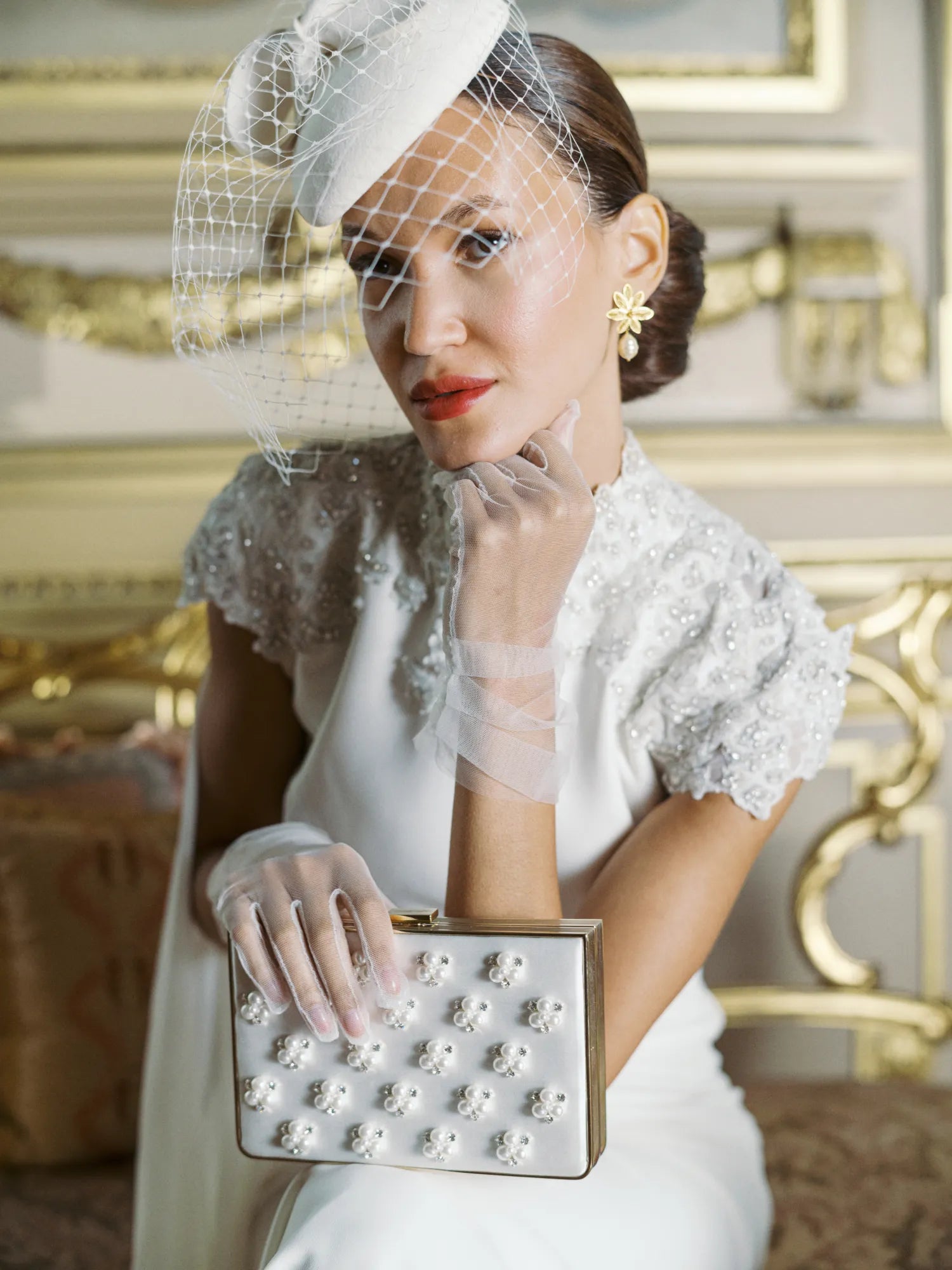 Woman in wedding dress &amp; wedding hat wearing tulle white gloves holding white satin clutch.