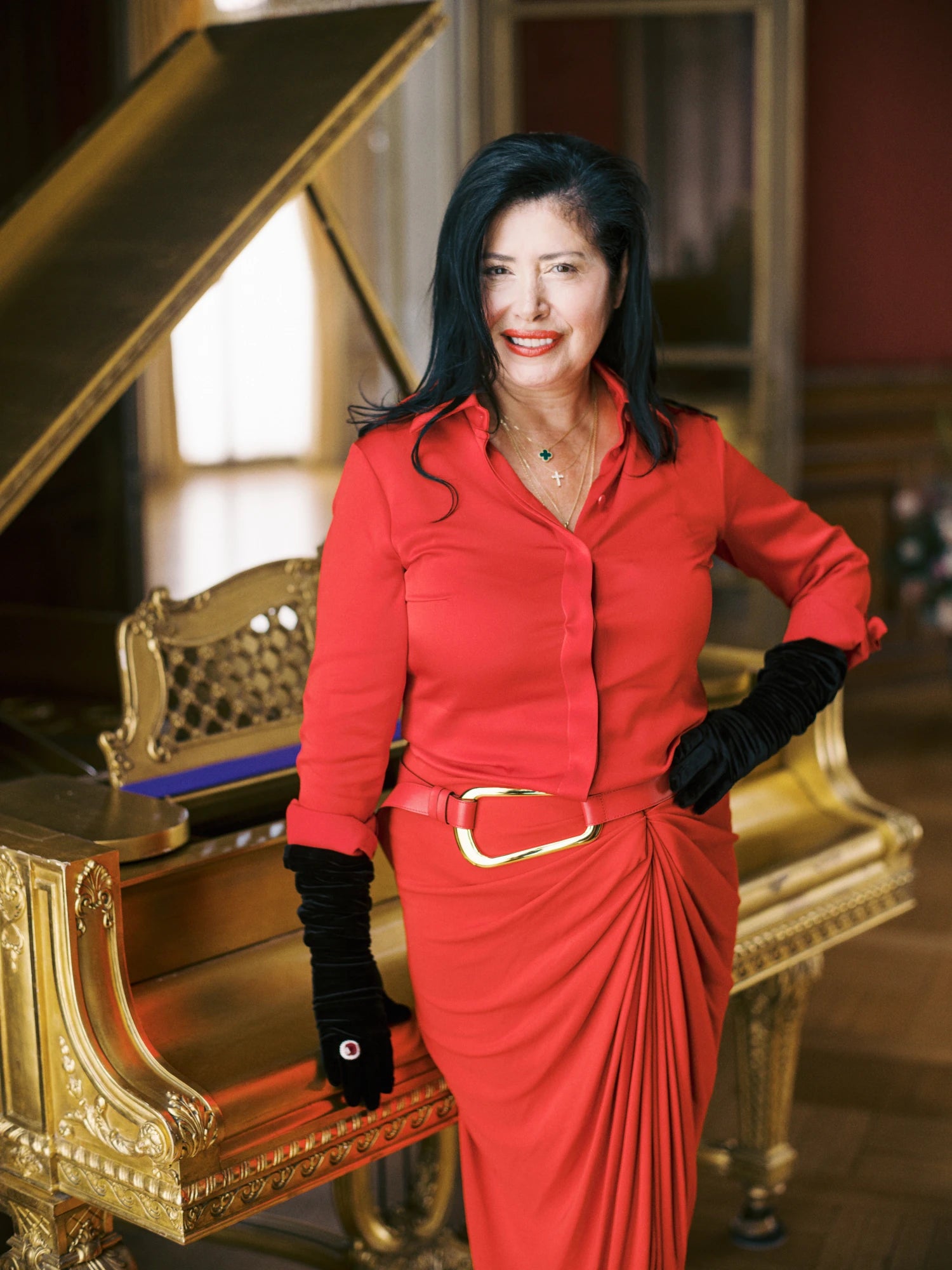 Woman wearing red dress and black gloves standing near gold grand piano.