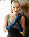 An elegant woman stands confident, wearing dark blue elbow length gloves.